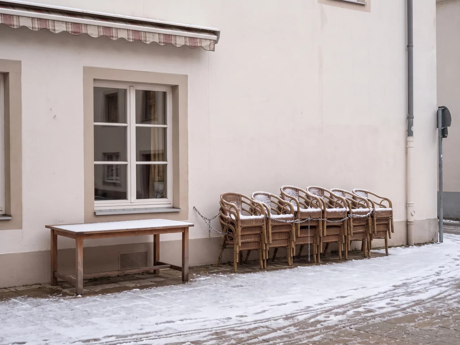 Dieselbe Terrasse im Winter: Markise eingefahren, Stühle gestapelt und angekettet, Schnee auf dem Pflaster